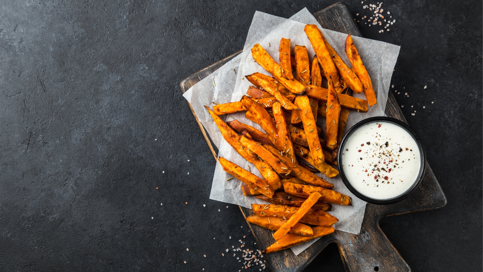 Oven-baked sweet potato fries seasoned with garlic and rosemary, served on parchment paper with a creamy dipping sauce.